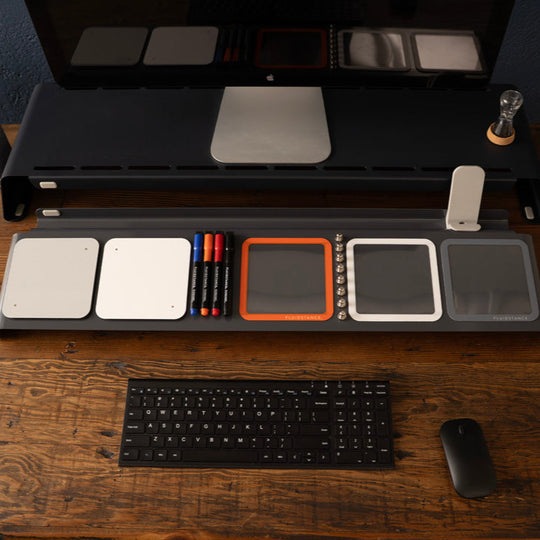 Dry erase Desk setup with a computer monitor riser, keyboard, mouse, and various stationery items on a wooden desk.
